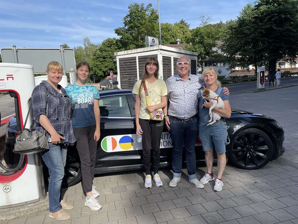 man from effective aid ukraine tesla convoy smiling with four smiling ukrainian refugees and dog in front of tesla car